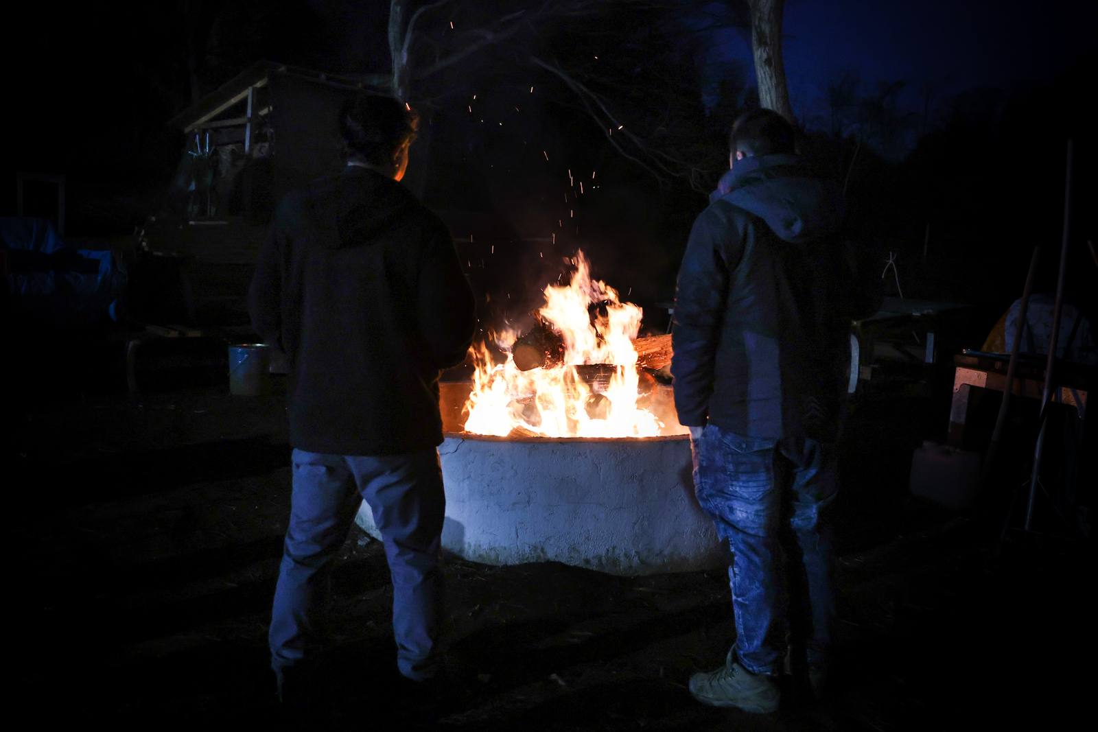 夜の里山で燃え上がる大焚き火。炎を囲む人の影が浮かんでいる。